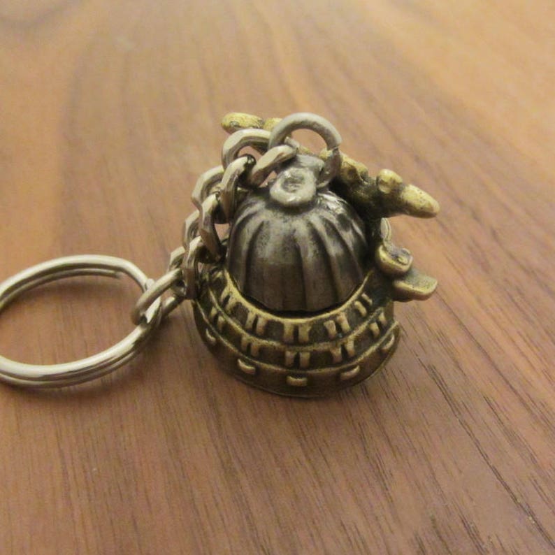 May include: A detailed keychain featuring a miniature helmet design. The helmet is made of metal with a silver and gold finish, attached to a chain and keyring. The helmet has intricate detailing and a vintage aesthetic.