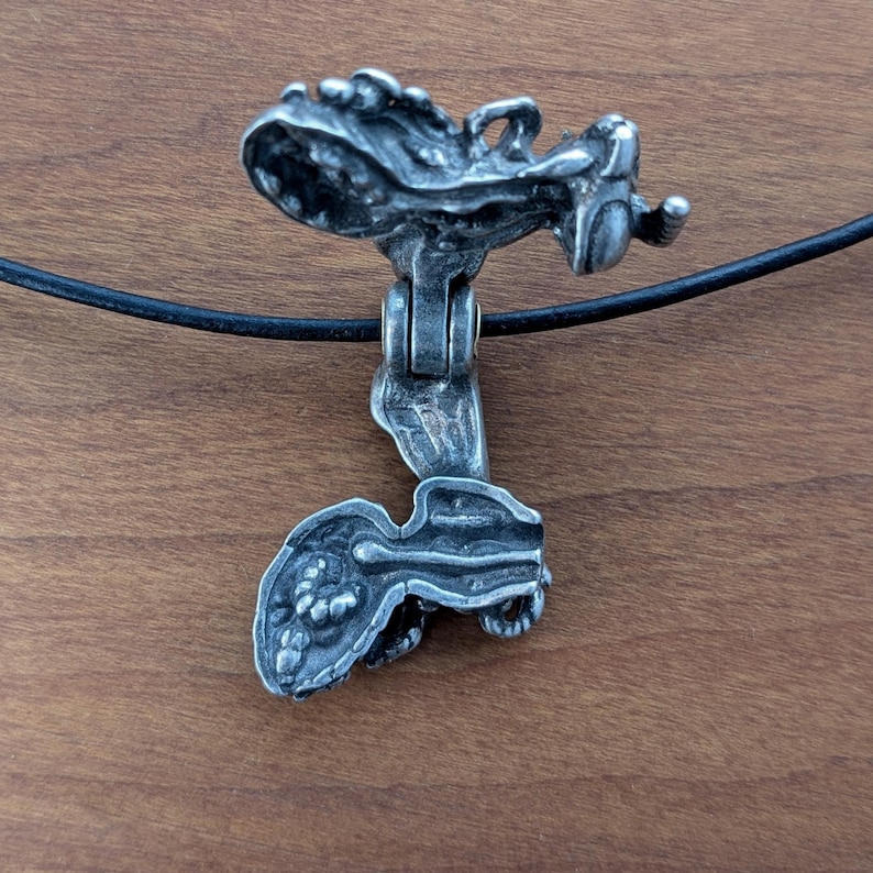 May include: A silver pendant necklace with a detailed, textured design resembling a creature. The pendant is open and has a hinged opening. The necklace is on a black cord.
