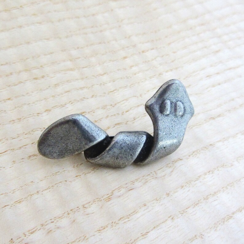 May include: A silver-toned metal drawer pull with a twisted design. The pull features a decorative head with two small, circular details. The hardware is shown against a light wood grain background.