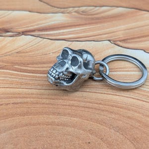 Puede incluir: Un llavero de metal plateado con un dije detallado en forma de cr&aacute;neo de primate. El cr&aacute;neo tiene arcos superciliares prominentes, una mand&iacute;bula definida con dientes visibles y un acabado liso y desgastado. Incluye una argolla met&aacute;lica.