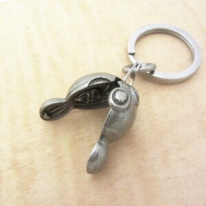 May include: A silver-colored metal keychain featuring a detailed, open clam shell design. The shell reveals an interior design. The keychain includes a split ring for attaching keys.