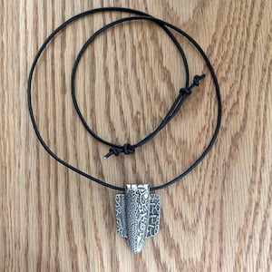 Puede incluir: Collar con colgante plateado y cord&oacute;n negro. El colgante presenta un dise&ntilde;o texturizado con una forma ovalada central y dos paneles laterales. El cord&oacute;n es ajustable con un nudo corredizo. El collar se exhibe sobre una superficie de madera.