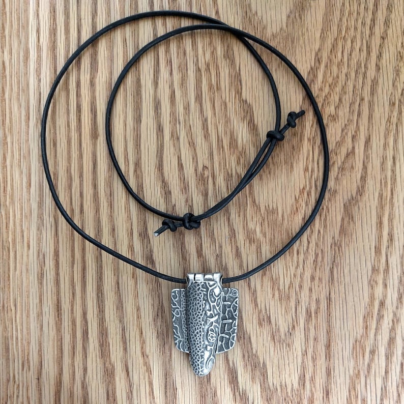 Puede incluir: Collar con colgante plateado y cord&oacute;n negro. El colgante presenta un dise&ntilde;o texturizado con una forma ovalada central y dos paneles laterales. El cord&oacute;n es ajustable con un nudo corredizo. El collar se exhibe sobre una superficie de madera.