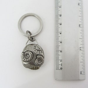 May include: A silver-colored keychain with a fossil-shaped pendant. The pendant features three spiral designs and a textured surface. The keychain includes a metal ring and a small connecting loop. The ruler next to the keychain measures in inches.