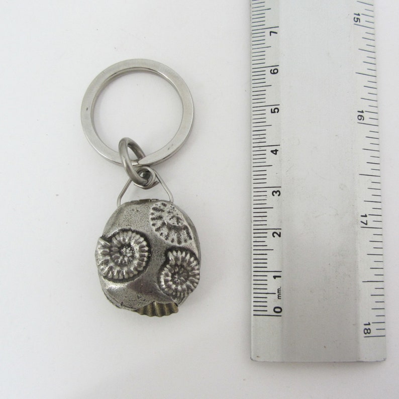 May include: A silver-colored keychain with a fossil-shaped pendant. The pendant features three spiral designs and a textured surface. The keychain includes a metal ring and a small connecting loop. The ruler next to the keychain measures in inches.