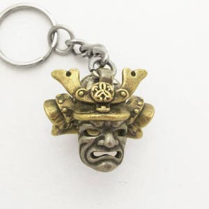May include: A detailed keychain featuring a samurai mask pendant. The pendant is a combination of gold and silver tones, with intricate detailing on the helmet and face. The keychain includes a silver-colored ring and chain.