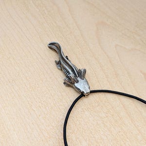 May include: A silver-colored axolotl pendant on a black cord. The pendant is detailed with visible gills and a textured body. The axolotl is a salamander known for its unique appearance. The cord is a simple, thin black band.
