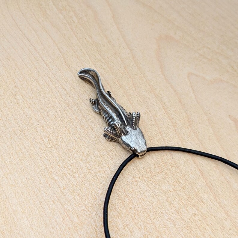 May include: A silver-colored axolotl pendant on a black cord. The pendant is detailed with visible gills and a textured body. The axolotl is a salamander known for its unique appearance. The cord is a simple, thin black band.