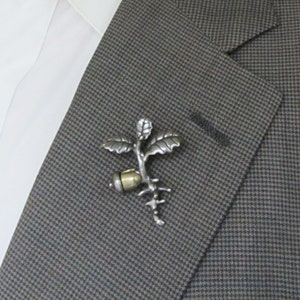 May include: A silver-toned metal lapel pin in the shape of a stylized plant with leaves, a seed, and roots. The pin is attached to a gray and brown patterned blazer, with a white shirt visible in the background.