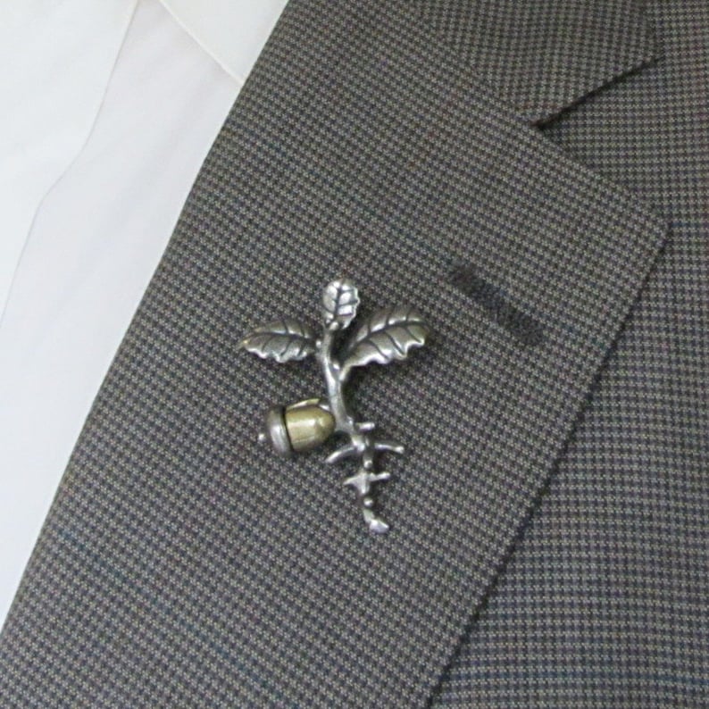 May include: A silver-toned metal lapel pin in the shape of a stylized plant with leaves, a seed, and roots. The pin is attached to a gray and brown patterned blazer, with a white shirt visible in the background.