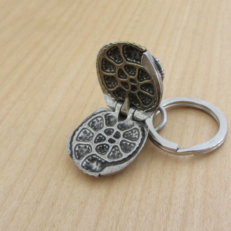 May include: A silver-colored, hinged keychain pendant with a detailed, open design. The pendant is attached to a silver key ring. The open pendant reveals an intricate pattern. The background is a light brown wooden surface.