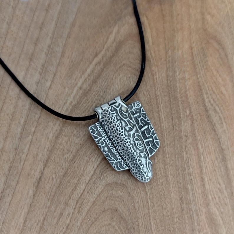 Puede incluir: Un colgante plateado con un dise&ntilde;o abstracto, suspendido de un cord&oacute;n negro. El colgante presenta patrones y formas texturizadas, incluyendo una secci&oacute;n central con una textura punteada y paneles laterales con dise&ntilde;os arremolinados. El collar descansa sobre una superficie de madera.