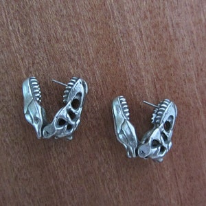 May include: A pair of silver-colored earrings shaped like dinosaur jaws. Each earring features detailed teeth and a skeletal design, with a post for pierced ears. The earrings are displayed on a wooden surface.