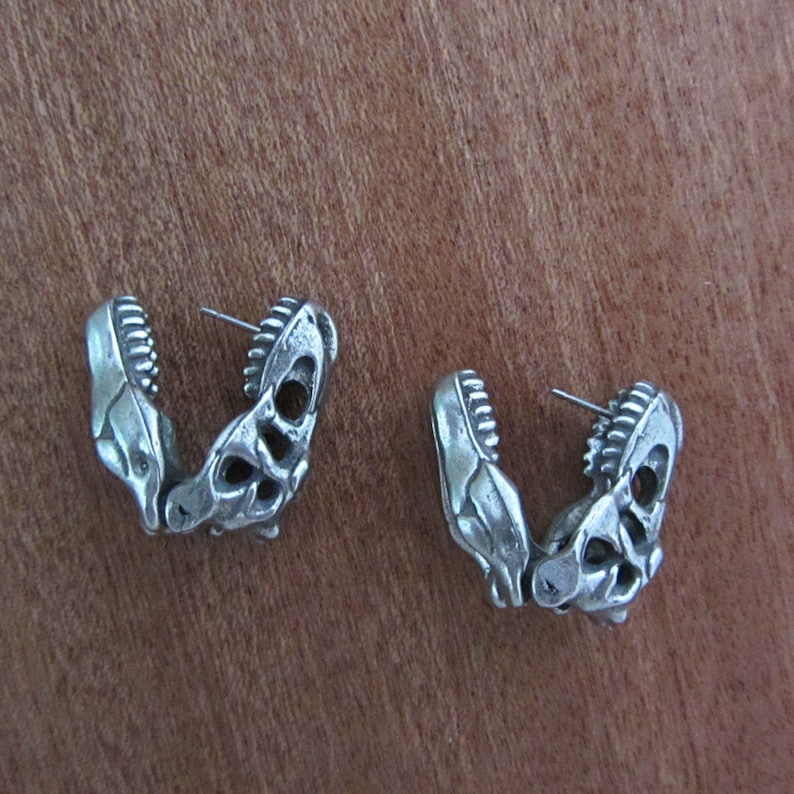 May include: A pair of silver-colored earrings shaped like dinosaur jaws. Each earring features detailed teeth and a skeletal design, with a post for pierced ears. The earrings are displayed on a wooden surface.