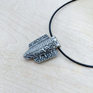 Puede incluir: Un collar con colgante plateado con un dise&ntilde;o texturizado y detallado. El colgante, con forma de pez o reptil estilizado, cuelga de un cord&oacute;n negro. Los intrincados patrones y el contraste crean un efecto visual llamativo.