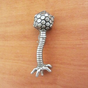 May include: A silver-toned brooch shaped like a stylized virus with a hexagonal head and a segmented body ending in a three-pronged base.