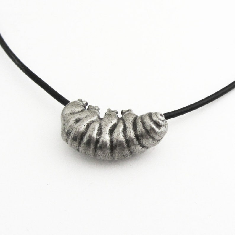 May include: A silver-toned caterpillar pendant necklace. The caterpillar has detailed ridges and is strung on a black cord. The necklace is set against a white background.