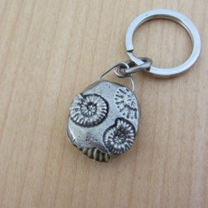 May include: A silver-colored keychain featuring a fossil design. The fossil pendant has three spiral-shaped imprints and is attached to a metal keyring. The keychain is a unique accessory.