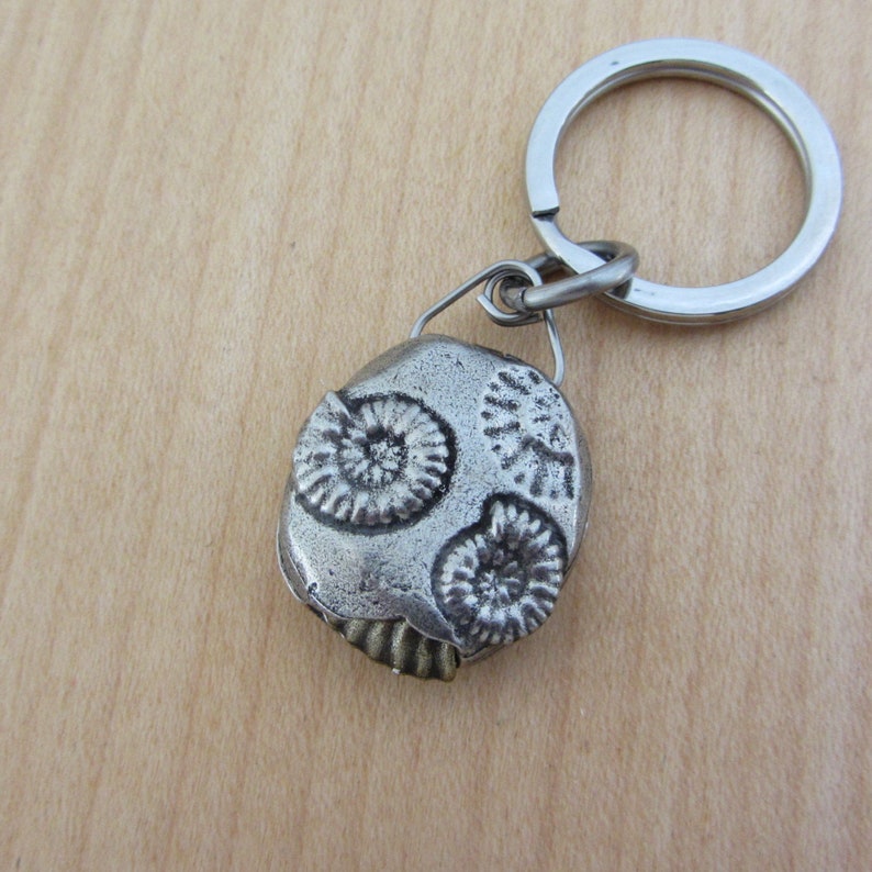 May include: A silver-colored keychain featuring a fossil design. The fossil pendant has three spiral-shaped imprints and is attached to a metal keyring. The keychain is a unique accessory.