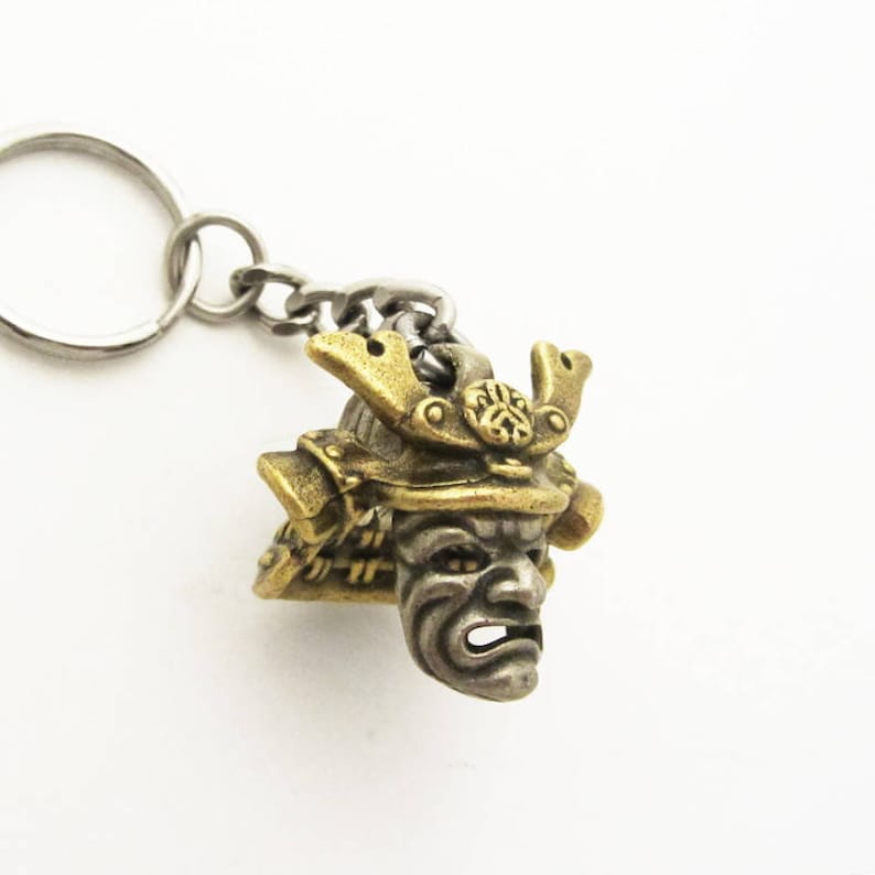 May include: A bronze-colored metal keychain featuring a detailed miniature samurai mask. The mask has intricate details, including a helmet and facial features. The keychain includes a silver-colored chain and key ring.