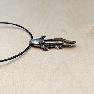 May include: A silver axolotl pendant on a black cord necklace. The detailed pendant features the salamander's distinctive external gills and a long, flattened tail. The necklace is displayed on a light wood surface.