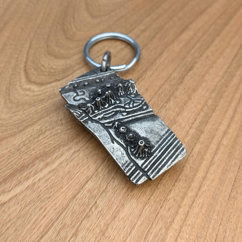 May include: A silver-colored metal keychain with a rectangular pendant. The pendant features a detailed landscape design with mountains, trees, and a textured surface. The keychain has a silver ring for attaching keys.