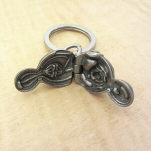 May include: A pewter-colored, hinged keychain pendant with a Celtic knot design. The pendant is open, revealing the interior details. A silver-colored keyring is attached to the pendant.
