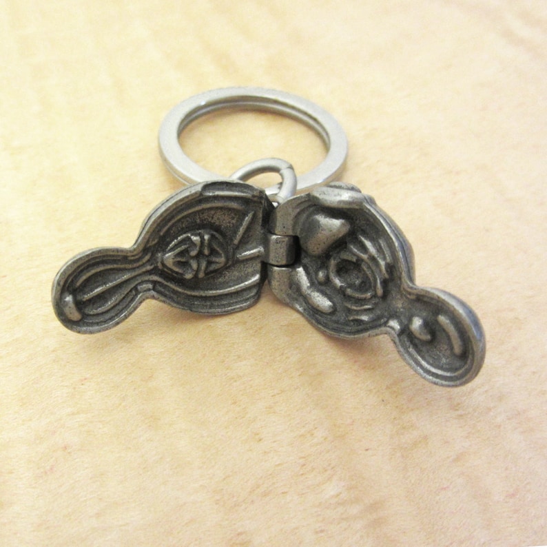 May include: A pewter-colored, hinged keychain pendant with a Celtic knot design. The pendant is open, revealing the interior details. A silver-colored keyring is attached to the pendant.