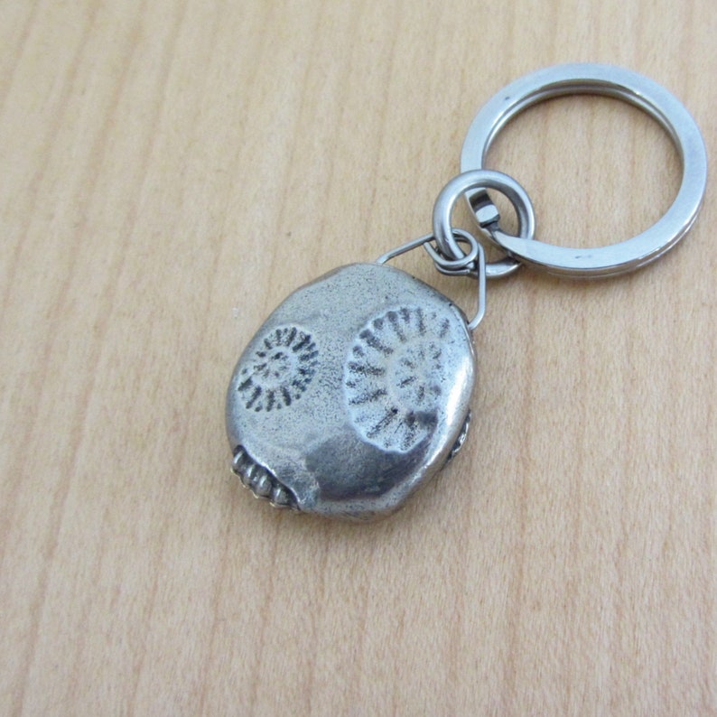 May include: A silver-colored keychain with a fossil-shaped pendant. The pendant features two spiral designs and a textured surface. The keychain ring is made of shiny metal, and the background is a light wood grain.