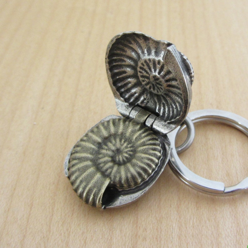 May include: A silver-colored, hinged keychain pendant shaped like an ammonite fossil. The pendant opens to reveal the fossil's spiral pattern. A silver key ring is attached.