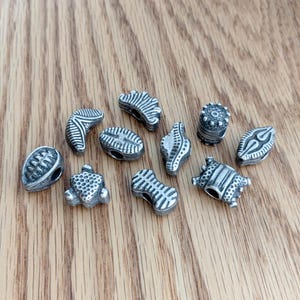May include: A collection of ten small, silver-toned metal beads with various embossed geometric patterns, including lines, dots, and ridges. Each bead features a central hole for threading, showcasing a weathered, antiqued finish on the surface.