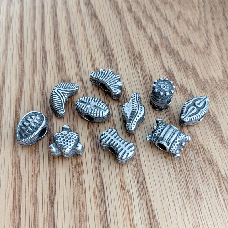 May include: A collection of ten small, silver-toned metal beads with various embossed geometric patterns, including lines, dots, and ridges. Each bead features a central hole for threading, showcasing a weathered, antiqued finish on the surface.