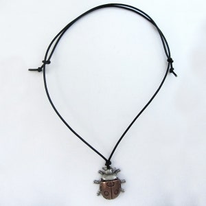 May include: A necklace featuring a metal ladybug pendant with silver and copper tones, suspended from a black cord. The ladybug has detailed wings and body segments. The cord is adjustable.