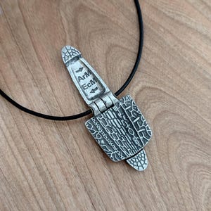 Puede incluir: Un collar con colgante plateado con un cord&oacute;n negro. El colgante tiene forma rectangular con patrones intrincados y el texto "ArM EcM". El colgante est&aacute; hecho de metal plateado y tiene un dise&ntilde;o articulado.