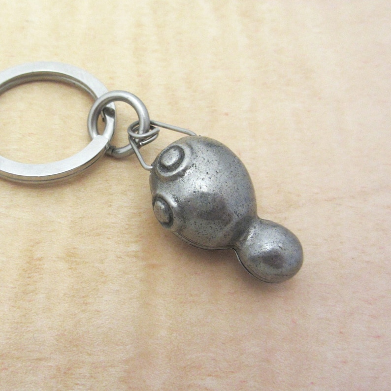 May include: A silver-colored metal keychain featuring a stylized tadpole-shaped charm. The charm has a textured surface and two circular details on the head. The keychain includes a split ring and a smaller connecting ring.