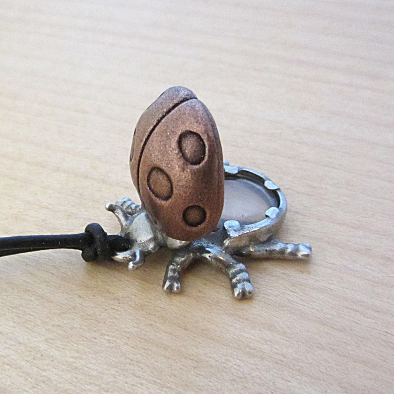 May include: A ladybug pendant with a copper-colored body and a silver-colored base. The ladybug has three circular spots on each side. The pendant is attached to a black cord.