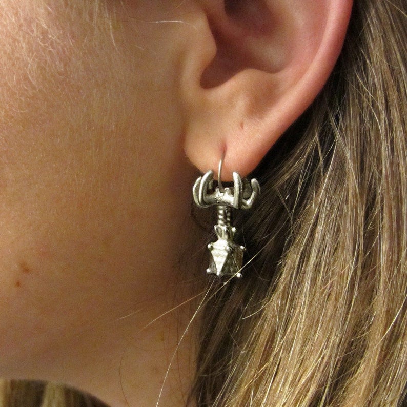 May include: Silver earrings with a geometric design. The earrings are shaped like a small, multi-faceted crystal with a small, skeletal figure attached to the top.