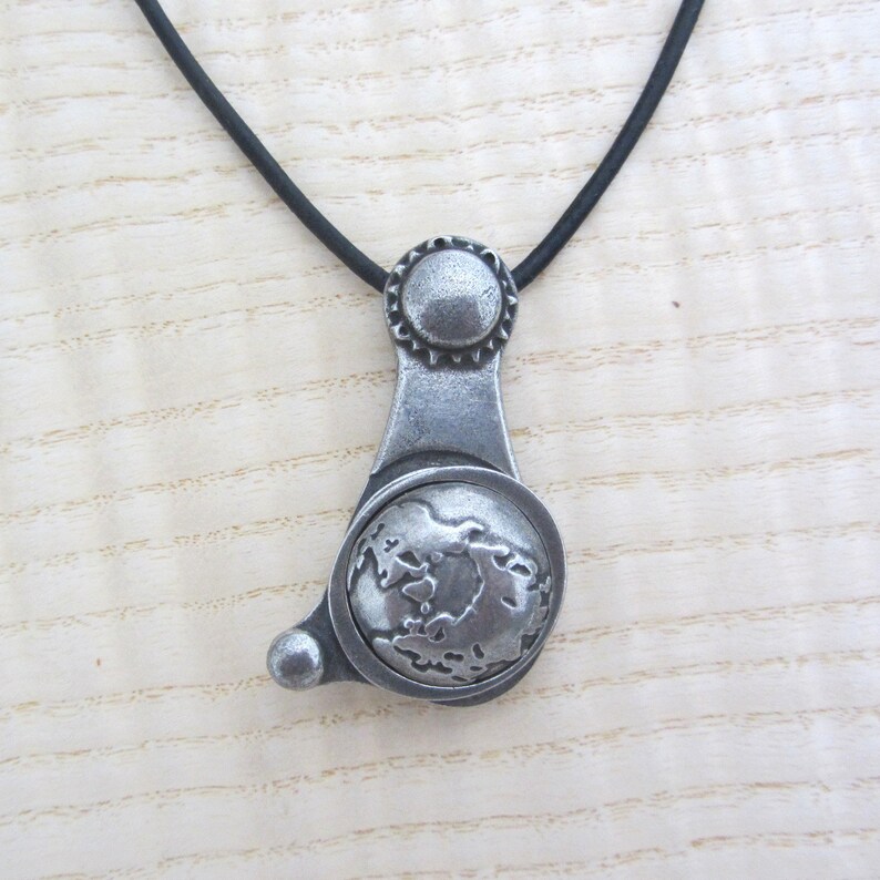 May include: A silver necklace with a pendant shaped like a globe. The pendant has a textured surface and a raised design of the continents. The necklace is on a black cord.