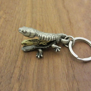 May include: A silver metal lizard-shaped keyring. The design features a larger lizard over a smaller gold-coloured lizard. The keyring has a textured finish and a silver key ring attached.