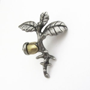 May include: A silver-toned metal brooch in the shape of a small branch with leaves and an acorn. The acorn has a gold-colored cap. The brooch is detailed with textured surfaces and is set against a white background.