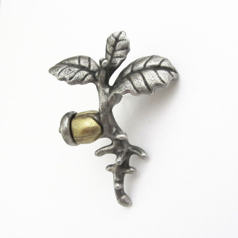 May include: A silver-toned metal brooch in the shape of a small branch with leaves and an acorn. The acorn has a gold-colored cap. The brooch is detailed with textured surfaces and is set against a white background.
