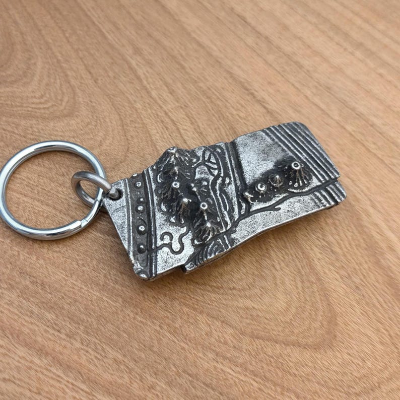 May include: A silver-colored metal keychain with an ornate rectangular pendant. The pendant features a detailed relief design with floral and abstract elements. The keychain ring is circular and attached to the pendant.
