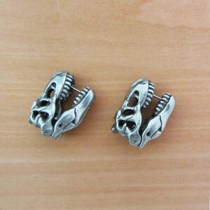 May include: A pair of silver-colored dinosaur skull stud earrings. Each earring is a detailed miniature of a T-Rex skull, with visible teeth and eye sockets. The earrings are set against a light brown wooden surface.
