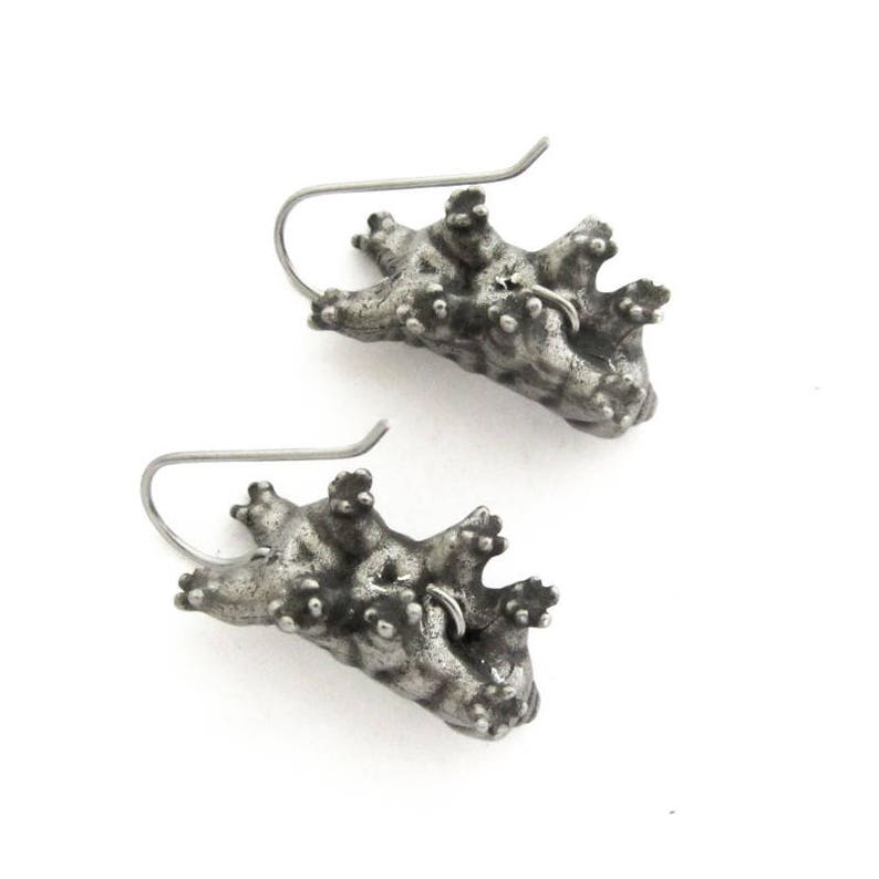 May include: A pair of silver-toned earrings shaped like tardigrades, also known as water bears. Each earring features a textured, segmented body with multiple small legs and a curved metal ear wire for attachment, showcasing a dark, oxidized finish.