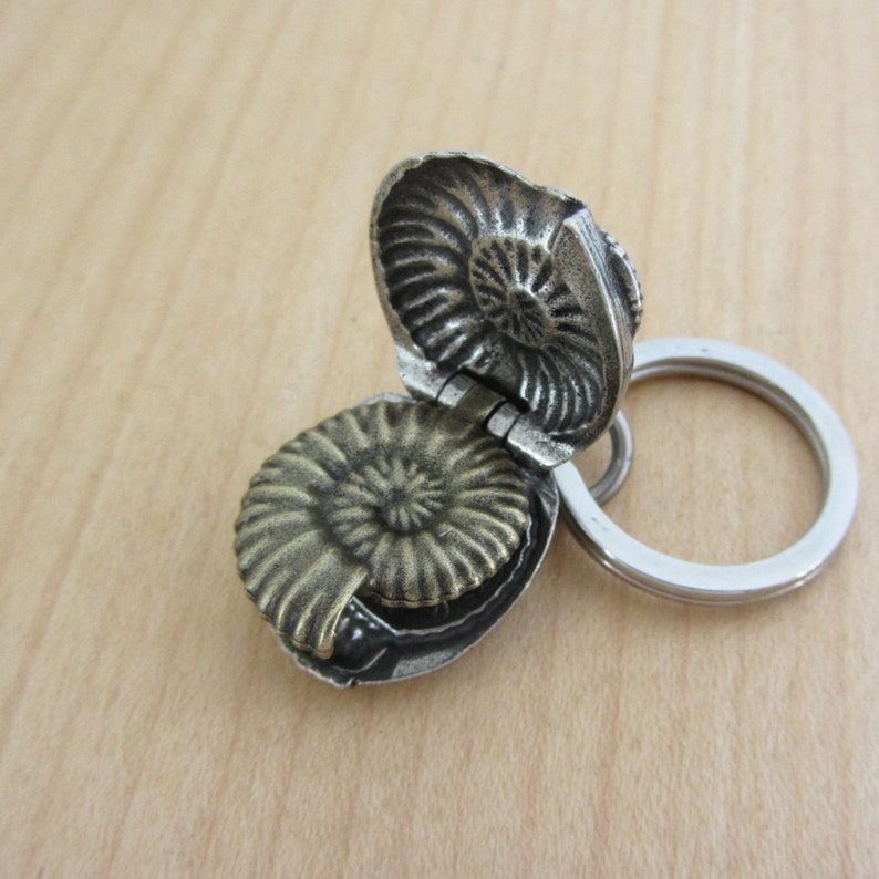 May include: A silver-toned, hinged keychain pendant shaped like an ammonite fossil. The pendant opens to reveal a detailed fossil design inside. A silver key ring is attached.