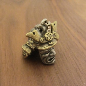 May include: A small, detailed bronze-colored miniature samurai helmet. The helmet features intricate designs, including a face mask, decorative plates, and a crest. The overall aesthetic is antique and detailed, suggesting a collectible or decorative item.