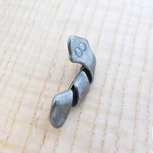 May include: A silver-toned metal brooch with a twisted, abstract design. The brooch has a pin closure on the back and features a small, rounded head with two indented circles. The overall shape is curved and elongated.