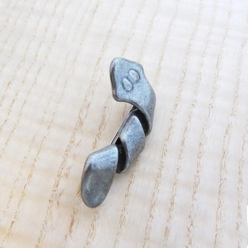 May include: A silver-toned metal brooch with a twisted, abstract design. The brooch has a pin closure on the back and features a small, rounded head with two indented circles. The overall shape is curved and elongated.
