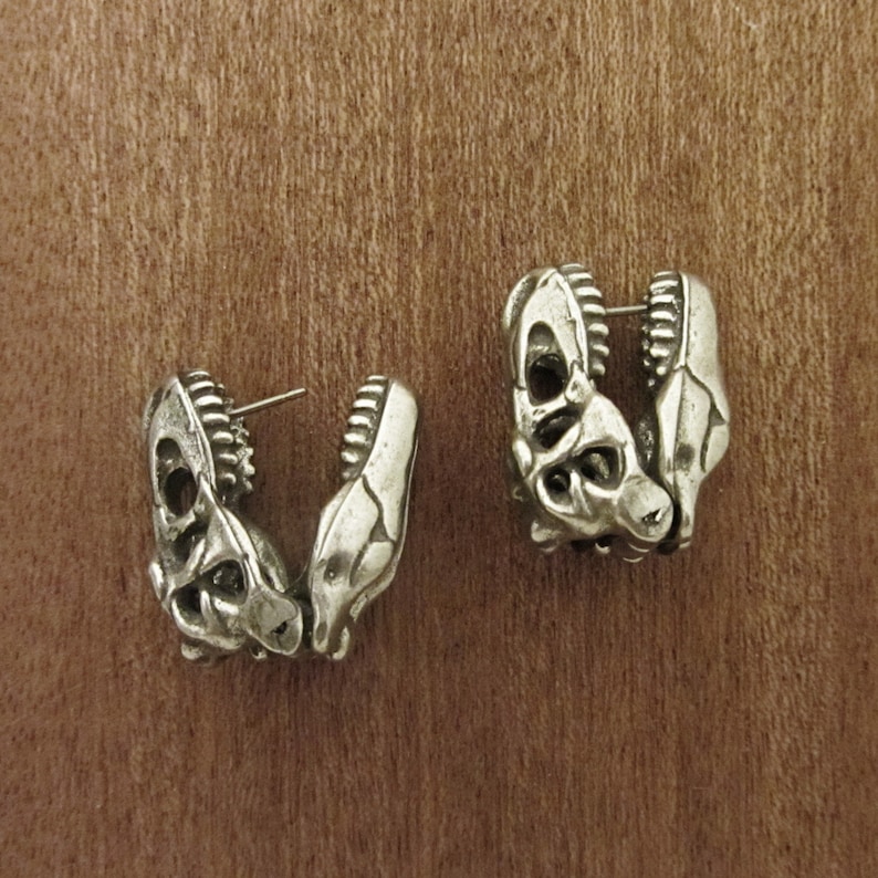 May include: A pair of silver-toned earrings shaped like dinosaur skulls. Each earring features detailed skeletal structures, including teeth and eye sockets. The earrings are displayed on a wooden surface, showcasing their intricate design.
