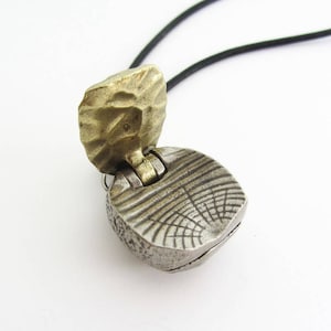 May include: A silver and brass pendant necklace with a hinged, textured, round locket. The locket is open, revealing the inside.